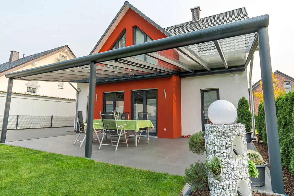 Gläserne Terrassenüberdachung aus Aluminium an einem roten Wohnhaus mit weitläufigem Garten